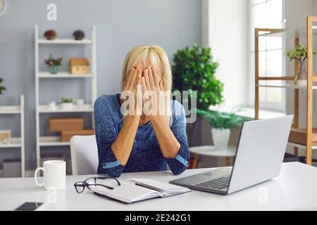 Donna anziana sconvolta seduta alla scrivania con computer portatile e copertura la faccia non è in grado di risolvere il problema Foto Stock