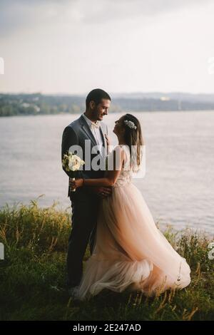 Immagine emozionale di coppia appena sposata in piedi sul campo e guardarsi l'un l'altro con amore. Fiume sullo sfondo. Foto Stock