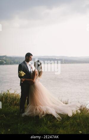 Immagine emozionale di coppia appena sposata in piedi sul campo e guardarsi l'un l'altro con amore. Fiume sullo sfondo. Foto Stock