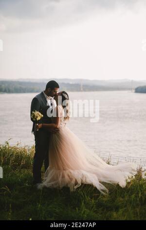 Immagine emozionale di coppia appena sposata in piedi in campo e baciando. Fiume in background.Couple gol. Foto Stock