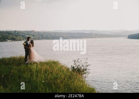 Immagine emozionale di coppia appena sposata in piedi in campo e baciando. Fiume in background.Couple gol. Foto Stock