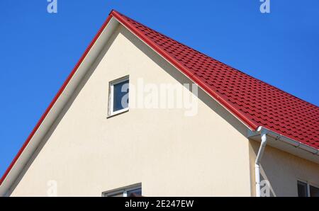 Un primo piano su un tetto in metallo rosso e soffito di plastica di una casa con pareti in stucco, piccola finestra soffitta e sistema di gocciolatoio tetto. Foto Stock