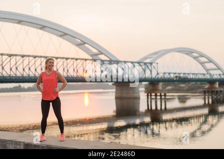 Ragazza bionda sportiva forte e sicura in piedi sul molo vicino al fiume dopo l'allenamento della mattina presto. Foto Stock