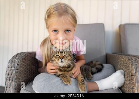 Carino adorabile bionda caucasica dolce bambina otto anni godere seduta in poltrona all'aperto tenendo e abbracciando piccolo gatto pappagallo in mani. PET Foto Stock