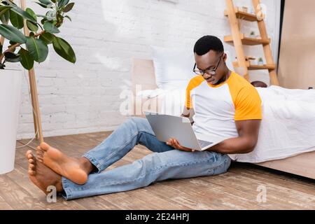 Freelance afroamericana a piedi nudi che usa il computer portatile mentre si siede vicino al letto e pianta a casa Foto Stock