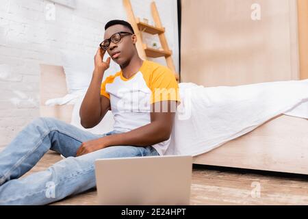 Uomo afro-americano stanco in occhiali seduto vicino al computer portatile su primo piano sfocato nella camera da letto Foto Stock