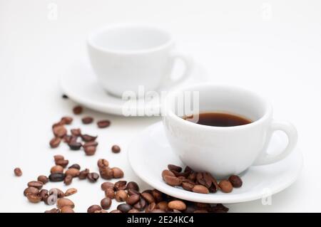 Due tazze per cappuccino bianche con piattini e chicchi di caffè tostati sparsi su sfondo bianco. L'attenzione è rivolta alla tazza anteriore riempita di bevanda a base di caffè. Foto Stock