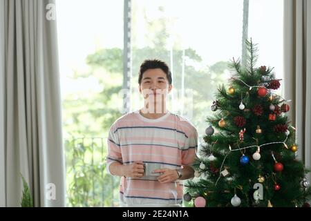 Giovane con tazza da caffè come regalo per le feste con decorate albero in background Foto Stock