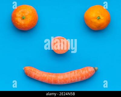 Una divertente composizione di mandarini e carote nella forma di un volto sorridente su uno sfondo azzurro Foto Stock