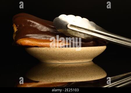 Un eclair al cioccolato con piattino in ceramica e pinze per pasticceria, su sfondo nero. Foto Stock