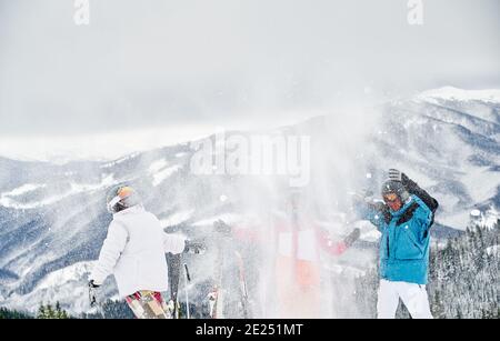 Amici sciatori in tute da sci gettando neve fresca in alto nell'aria. Due uomini e una donna che si divertono su una collina innevata con bellissime montagne sullo sfondo. Concetto di intrattenimento invernale, amicizia. Foto Stock