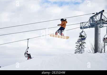 Snowboarder fare trucchi in montagna. Ragazzo che vola in alto con snowboard contro lo skilift e il cielo nuvoloso. Vista ad angolo basso. Concetto di sport invernali, sport estremi nella stazione sciistica Foto Stock