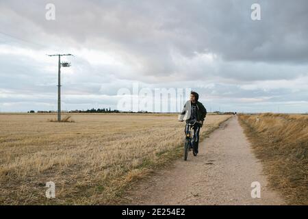Giovane uomo che guida la sua bici d'epoca in una strada sterrata accanto a un campo asciutto. Giorno nuvoloso con costume scheletro Foto Stock