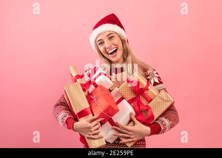 Ritratto di donna bionda felice in felicissimo maglione e Santa cappello che tiene un mucchio di regali di Natale su sfondo rosa Foto Stock