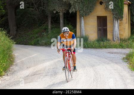 Ciclista in bicicletta passando accanto alla chiesetta e ai cipressi di Lucignano d'Asso, partecipando a maggio all'Eroica Montalcino, Siena, Toscana, Italia Foto Stock