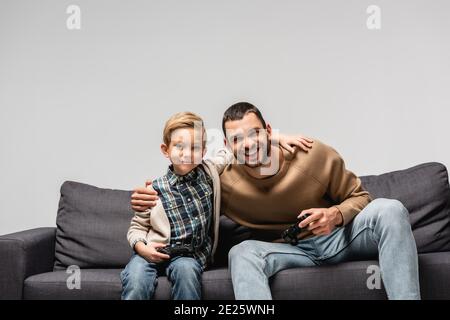 KIEV, UCRAINA - 17 NOVEMBRE 2020: Felice padre e figlio abbracciare mentre tenendo i joystick isolati su grigio Foto Stock