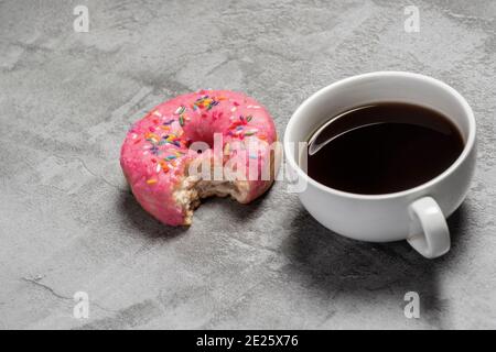 Ciambella smaltata rosa con spruzzi e un morso preso di esso accanto a una tazza nera di caffè con una vista angolata Foto Stock