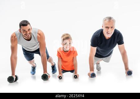 ragazzo allegro con nonno e papà che fanno esercitare spinta su con manubri su bianco Foto Stock