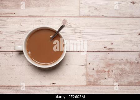 Tazza di tè o caffè forte con latte e un cucchiaio in una tazza bianca su una superficie testurizzata di legno bianco con spazio per la copia e spazio per il testo. Foto Stock