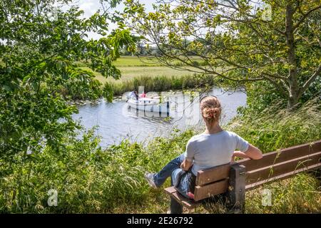 Donna seduta sulla panchina sulla riva del fiume guardando la barca da diporto sul fiume Linge vicino a Rhenoy, Betuwe occidentale, Gelderland, Paesi Bassi Foto Stock