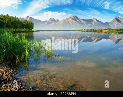 paesaggio composito di lago di montagna in estate. splendido paesaggio naturale in una mattina soleggiata. cielo e cresta che si riflettono nella calma superficie d'acqua. alberi Foto Stock