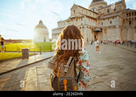Vista da dietro elegante donna turistica in abito floreale con zaino facendo escursioni vicino alla Cattedrale di Pisa. Foto Stock