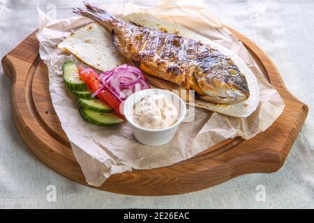 Pesce dorado alla griglia con salsa, cetrioli a fette, pomodori e cipolle su un vassoio di legno Foto Stock