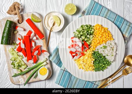 Insalata di granchio di bastoncini di surimi con mais in scatola, cetrioli, uova sode con cipolla primaverile, riso di gelsomino diviso in settori su un piatto bianco su un bianco Foto Stock