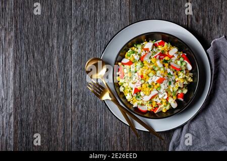 Insalata di granchio di granchio artificiale con verdure: Mais, cetrioli, scalogno, uova sode, riso di gelsomino servito in un piatto nero su un legno scuro Foto Stock