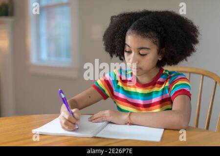 Dieci anni ragazza bi-razziale che scrive in notebook a tavola Foto Stock