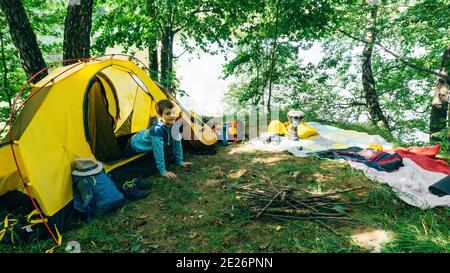 Un ragazzo si affaccia dalla tenda al mattino dopo aver trascorso la notte in un campo di famiglia sulla riva di un lago forestale. Un ragazzo carino è seduto in un giallo Foto Stock