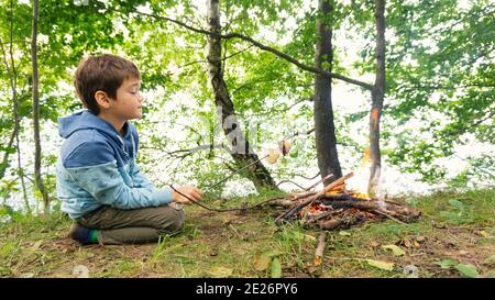 Il ragazzo fritta il pane su uno spiedino accanto al fuoco in un campo di famiglia. Lezioni di sopravvivenza nella foresta. Prima colazione nella foresta vicino al lago. Cucinare il cibo su un abete Foto Stock