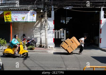 Bangkok, Thailandia - Dicembre, 2015: Uomo che maneggia il carrello con le scatole di cartone sul mercato di strada. Un altro uomo che cavalca la sua moto gialla sulla strada. Foto Stock