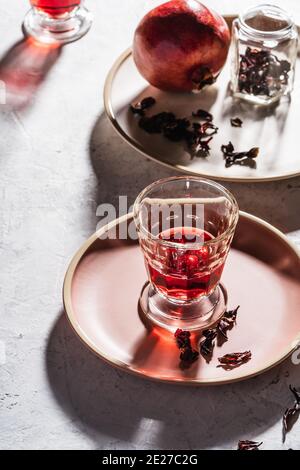 Rinfrescante tè alle erbe di frutta in bicchieri, melograno maturo su piatto, tè hibiscus e petali di rosa secco, luce dura con ombre dure Foto Stock