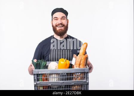 Foto di gioioso Chef man con barba che tiene una scatola con generi alimentari su sfondo bianco. Foto Stock