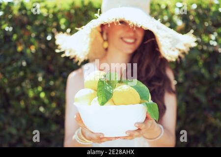 felice giovane casalinga in camicia bianca con piatto di limoni fattoria locale e cappello all'aperto vicino parete verde. Foto Stock