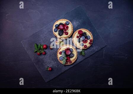 Vista dall'alto di tre torte con frutti di bosco e crema di vaniglia su un piatto di ardesia circondato da fragole. Foto Stock