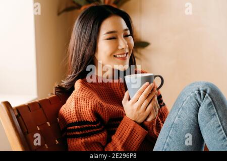 Felice giovane etnia femmina in maglione sorridente e godendosi caldo bevande mentre si riposa su una comoda sedia a casa Foto Stock