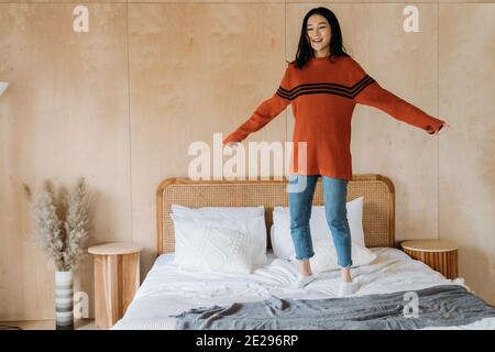 Allegra e rilassante donna etnica asiatica in maglione sorridente e saltellante a letto mentre ci si diverte a casa Foto Stock