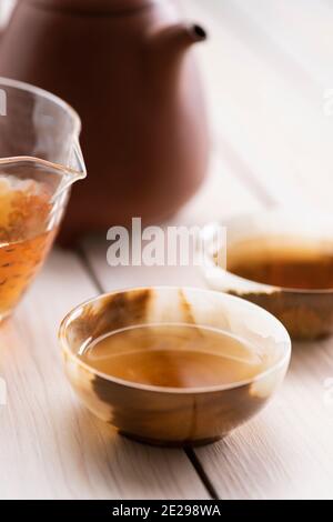 Primo piano su bevanda sana. Tè cinese vecchio shen puerh fatto in teiera di argilla e servito in chahai di vetro e tazze di pietra onice su Clo tavolo di legno bianco Foto Stock