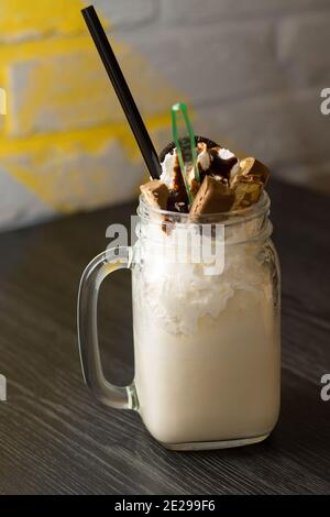Frullato in vaso con cannuccia da cocktail, pezzi di cioccolato e biscotti su tavolo di legno scuro Foto Stock