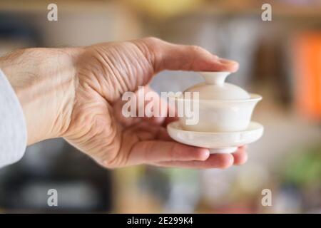 Mano dell'uomo che tiene il gaiwan bianco piccolo, tazza cinese di porcellana per la preparazione del tè primo piano Foto Stock