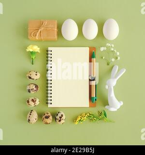 Buon concetto di festa di pasqua. Layout pasquale fatto di varie uova, coniglietto figurine, fiori, artigianato regalo e aprire blocco note vuoto su sfondo verde Top v Foto Stock