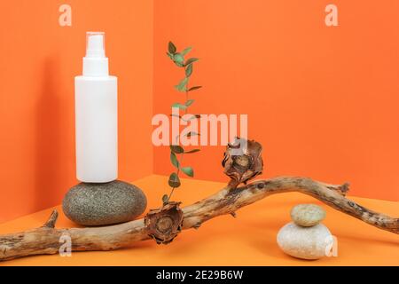Una bottiglia cosmetica bianca vuota con prodotto cosmetico su roccia, bastone di legno con fiori secchi e ramo di eucalipto nello spazio angolare sul backgro arancione Foto Stock
