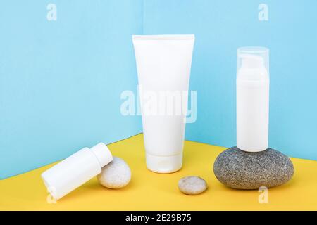 Bottiglie bianche per cosmetici in bianco, tubetto di crema, lozione per il corpo, il viso, la mano o altri prodotti cosmetici e rocce grigie su sfondo blu giallo. Mockup F Foto Stock