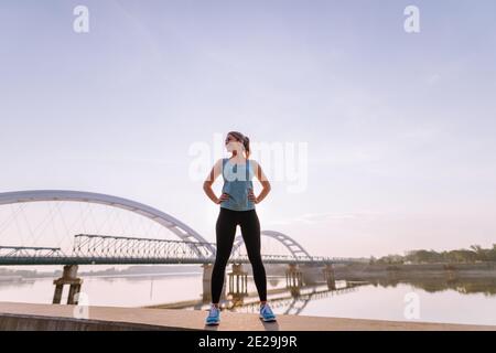 Ragazza bionda sportiva forte e sicura in piedi sul molo vicino al fiume dopo l'allenamento della mattina presto. Foto Stock