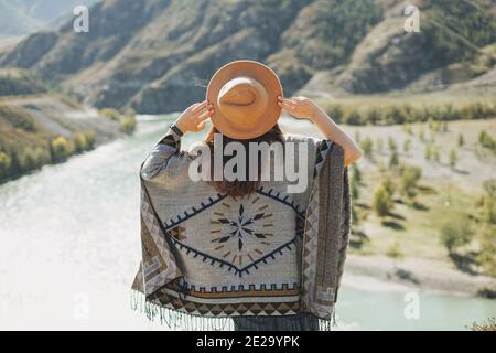 Spensierata bruna giovane donna viaggiatore in poncho da dietro sfondo del fiume di montagna Foto Stock