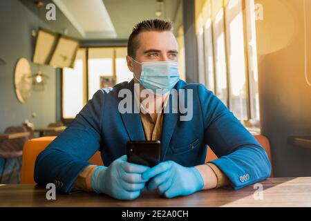 Giovane uomo d'affari europeo in guanti protettivi e una maschera medica passa il tempo sul telefono e guarda fuori la finestra. Foto Stock