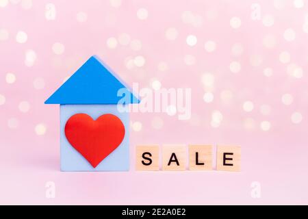 Legno blu casa con cuore rosso e con testo Vendita su sfondo bokeh rosa con copy-space. Noleggio di concetto, costruzione e acquisto domestico, compri un Foto Stock