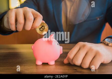 La mano di un uomo d'affari tiene una moneta di criptovaluta d'oro e mette un maiale in una banca piggy. Bitcoin, criptovaluta e concetto di risparmio. Foto Stock